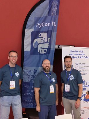 Three members of the PyCon NL team in matching Python t-shirts, standing beside the PyCon NL banner at their booth